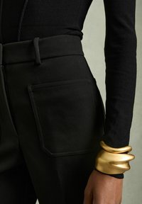 Primo piano di una persona che indossa pantaloni neri a vita alta con una grande tasca frontale e un braccialetto scultoreo dorato al polso.