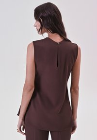 Donna con lunghi capelli scuri che indossa un top sans maniche di colore marrone scuro con un bottone sulla schiena e un dettaglio a forma di chiave, abbinato a pantaloni coordinati.