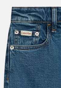 Jeans de mezclilla con un lavado azul, que presentan un bolsillo delantero con un remache plateado y una etiqueta blanca con "Calvin Klein" en letras rojas.