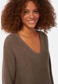 Pull en tricot marron avec un design en V, texture côtelée et coupe décontractée. Le tissu semble doux et confortable.