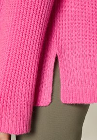 Maglione rosa acceso a coste con maniche lunghe e spacchi laterali. Il tessuto ha una consistenza morbida che aggiunge profondità al colore vibrante.