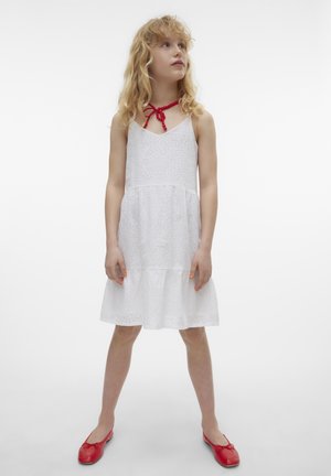 Robe en coton blanc avec des détails en broderie, corsage à volants et fines bretelles. Les accents rouges comprennent un collier et des chaussures assorties. Fond uni.