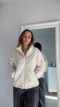 Kvinde iført en hvid fleecejakke og sorte bukser står foran et stort spejl, der reflekterer hendes ryg i et lyst rum.