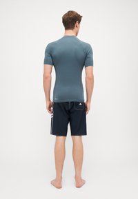 Menns tettsittende rashguard med kort erm i teal, kombinert med svarte badeshorts med hvite detaljer og en baklomme. Glatt tekstur.