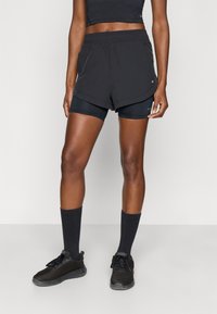 Pantalones cortos deportivos negros superpuestos sobre pantalones cortos de compresión negros, combinados con calcetines oscuros con costillas y zapatillas de malla negras. Textura suave.