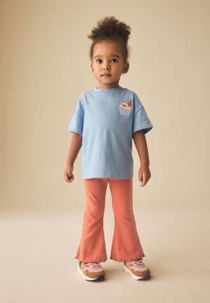 Malé dieťa s kučeravými vlasmi nosí svetlomodrú voľnú košeľu s nášivkou cupcake, koralové rozšírené nohavice a ružové tenisky, stojace na neutrálnom pozadí.