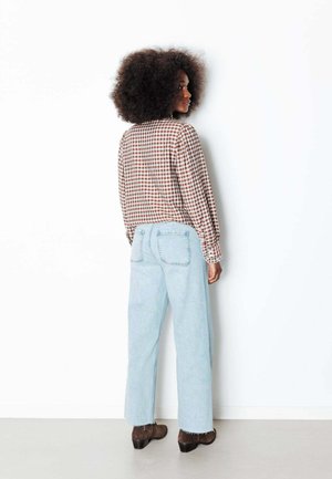 Femme aux cheveux bouclés portant une chemise à carreaux marron, un jean large bleu clair et des bottes marron, debout et regardant sur le côté.