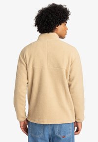 Beige fleece-tröja med hög krage och en mjuk, strukturerad yta. Har en bakre ok-söm och en avslappnad passform.