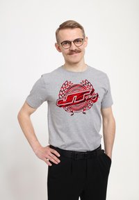 Šedé tričko s krátkým rukávem, zdobené červeným grafickým logem s vavřínovými akcenty, v kombinaci s černými kalhotami a černým opaskem.