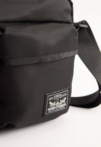 Borsa a spalla nera Levi Strauss & Co. con patch del marchio in evidenza e tracolla regolabile su sfondo bianco.