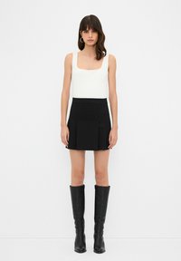 Jupe mini plissée noire taille haute, associée à un débardeur blanc à col carré ajusté et des bottes noires en cuir jusqu'aux genoux.