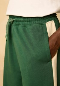 Zelené tepláky s elastickým pásom a šnúrkou na sťahovanie. Majú kontrastný béžový bočný pruh a hladkú, texturovanú tkaninu.