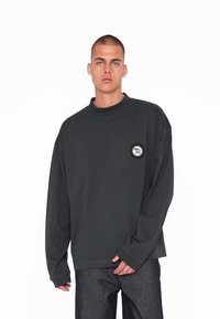 Sweatshirt supersize de culoare gri închis, cu guler înalt și mâneci lungi, având un logo rotund sub formă de patch pe partea stângă a pieptului.