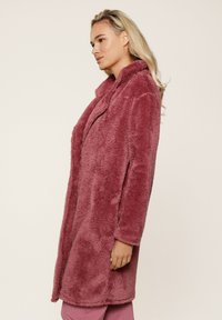 Cappotto in pelliccia sintetica di colore rosa tenue, con colletto, maniche lunghe e una silhouette dritta. La texture appare soffice e accogliente.