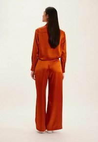 Ensemble deux pièces en satin orange avec manches longues et pantalons à jambes larges, présentant des poignets boutonnés et une texture lisse. Design visible au dos.