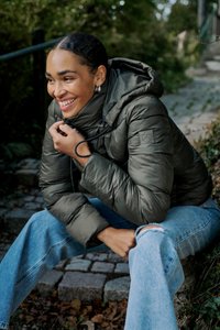 Ciemnozielona kurtka puchowa z kapturem, z zamkiem błyskawicznym z przodu i elastycznym sznurkiem. Nosi ją osoba w jasnoniebieskich jeansach z dziurami, siedząc na kamiennych schodach.