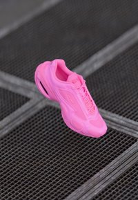Bright pink athletic sneaker with mesh and synthetic panels, floating above a metal grid floor with a crisscross pattern.