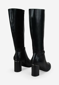 Botas altas de cuero negro con tacones de bloque y cremalleras laterales, mostradas desde una vista trasera en tres cuartos sobre un fondo blanco.
