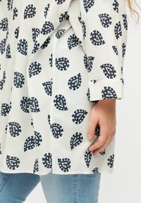 Άσπρο πουκάμισο με μακριά μανίκια και μαύρα σχέδια παϊένσι, φορεμένο με ανοιχτό μπλε τζιν, δείχνει μια τσέπη στην πλευρική ραφή και το χέρι ενός ατόμου να ακουμπά δίπλα της.