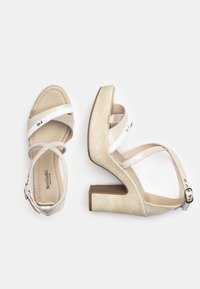 Beżowe lakierowane sandały na wysokim obcasie z krzyżującymi się paskami, teksturowany jasny słupkowy obcas oraz zapięcie na klamrę na kostce.