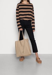 Beige canvas tote bag with rectangular logo, black and brown striped long-sleeve shirt, dark jeans, and brown platform sandals.