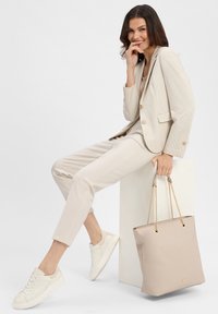 Beige maßgeschneiderter Anzug mit dezentem Hahnentrittmuster, kombiniert mit weißen Sneakers. Hält eine beige Shopper-Tasche mit goldenen Hardware-Akzenten.