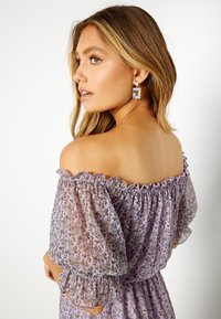 Jasnofioletowa sukienka odkrywająca ramiona z kwiecistym wzorem, falbaniastymi rękawami i teksturowanym materiałem. W komplecie prostokątne kolczyki z kamieniami szlachetnymi.