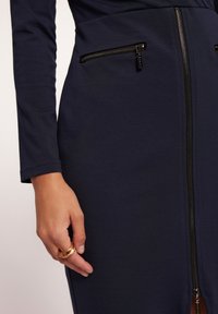 Granatowa dopasowana sukienka z długimi rękawami, z dwoma zamykanymi na zamek kieszeniami oraz centralnym zamkiem. Gładka tekstura i długość do połowy uda.