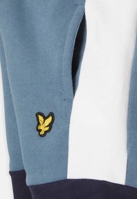 Pulôver feito de tecido suave, com um corpo azul, painéis brancos e detalhes em azul marinho. Um logo de águia bordado em amarelo está presente na manga.
