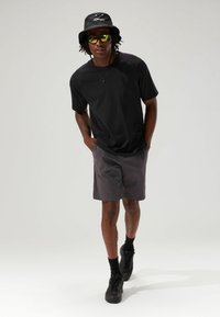 Schwarzes Kurzarmhemd mit lässiger Passform, kombiniert mit dunkelgrauen Shorts. Accessoires sind ein schwarzer Bucket Hat und reflektierende Sonnenbrille.