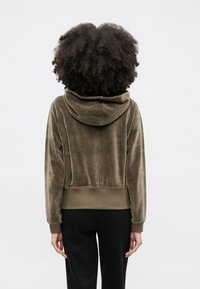 Pessoa com cabelo encaracolado vestindo um capuz de veludo verde-oliva e calças pretas, de pé frente a um fundo cinza claro e simples.