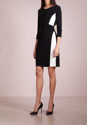 Woman wearing a knee-length black dress with white geometric panels, three-quarter sleeves, and black high-heeled shoes, standing on a plain floor.