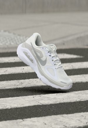 Chaussure de course Nike blanche et grise en l'air au-dessus d'un passage piéton sur l'asphalte, avec de petits débris volant en dessous.