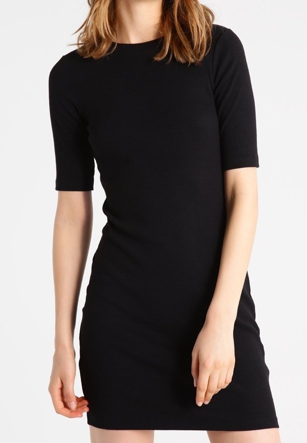 Femme portant une robe noire ajustée longueur genou avec des manches courtes, debout devant un fond blanc uni.