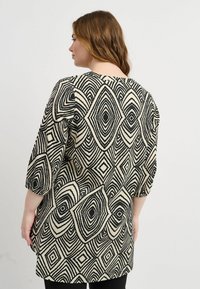 Schwarz-beige Tunika mit einem geometrischen Rautenmuster, Dreiviertelärmeln und lockerer Passform, aus einem weichen, strukturierten Stoff gefertigt.