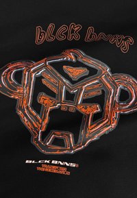 Zwarte shirt met een oranje grafiek van een gestileerd aapgezicht met geometrische accenten en vetgedrukte tekst, tegen een effen zwarte achtergrond.