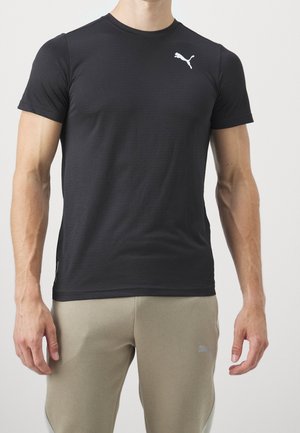 Mann trägt schlichtes schwarzes Puma-T-Shirt mit kurzen Ärmeln und beige Puma-Sweathosen, steht vor einem schlichten hellen Hintergrund.