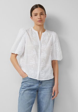 Frau mit braunen Haaren in einer weißen, mit Blumen bestickten Bluse mit Puffärmeln und hellblauen Jeans, die vor einer grauen Wand steht.