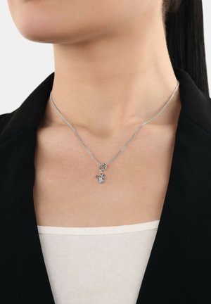 Silberne Kettenhalskette mit einem kleinen Engel-Anhänger, getragen von einer Person in einem schwarzen Blazer und einem weißen Oberteil, aus der Nähe fotografiert.