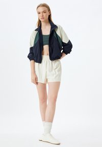 Tummansininen, vihreä ja kermaharmaa väriblokattu lyhyt takki, jossa on joustavat hihansuut, yhdistettynä vihreään lyhythihaiseen puseroon ja vaaleisiin kermaisiin shortseihin. Valkoiset lenkkarit mukana.