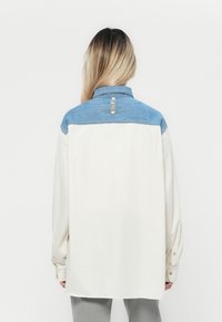 Person mit langen Haaren, die ein lockeres weißes Hemd mit einer blauen Passe und einem "REPLAY"-Label auf dem Rücken trägt, steht vor einem schlichten weißen Hintergrund.