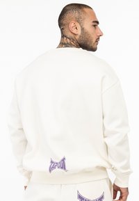 Hvid sweatshirt med lange ærmer, ribbede manchetter og et trykt lilla "TAPOUT" logo på ryggen. Glat tekstur, casual fit design.