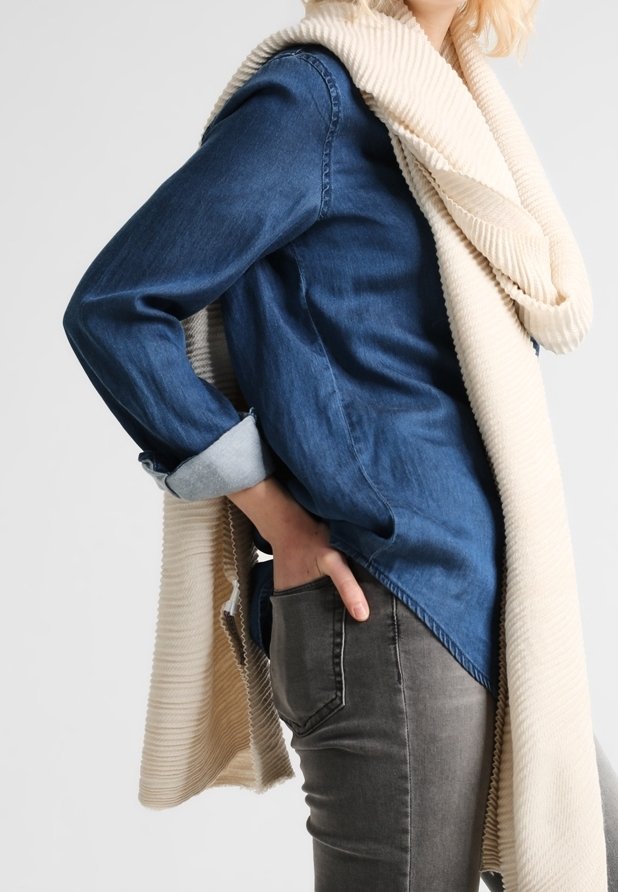 Personne portant une chemise en jean bleue avec les manches retroussées, un jean gris et une grande écharpe crème côtelée drapée sur les épaules.