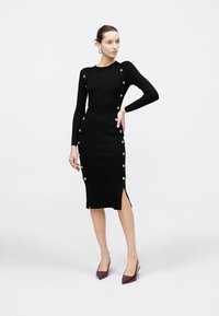 Czarna prążkowana dzianinowa sukienka z długimi rękawami, złotymi guzikami, rozcięciem z boku, dopasowaną sylwetką i długością do kolan.