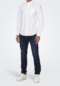 Valkoinen pitkähihaisen nappipaidan, jossa on rintatasku, yhdistetty tummansiniset slim fit -farkut ja valkoiset lenkkitossut, seisoo vaalealla taustalla.