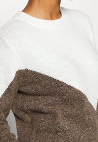 Tröjan har en vit övre del och en strukturerad brun nedre del, med en klassisk rund hals och mjukt stickat tyg.