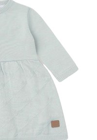 Vestido de bebé de punto azul claro con mangas largas, patrón texturizado de rombos en la falda y una pequeña etiqueta cuadrada marrón cerca del dobladillo.