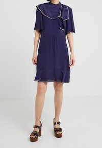 Mörkblå klänning med korta ärmar och lager-på-lager halsringning. Utsvängd nederdel med kontrasterande kant. Kombinerad med bruna platåskor.