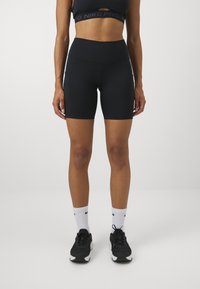 Kobieta w czarnych wysokich szortach sportowych, czarnym biustonoszu sportowym, białych skarpetach Nike i czarnych butach do biegania stoi na białym tle.