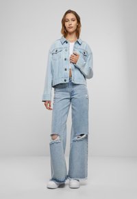 Chaqueta de mezclilla de color azul claro con un corte corto, cierre de botones y dos bolsillos delanteros. Combinada con jeans anchos deshilachados y zapatillas blancas.